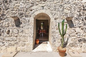 Ein Steinhaus mit einem Rundbogenportal, das zu einem Innenraum führt. Vor dem Portal stehen zwei Kakteen in Töpfen.