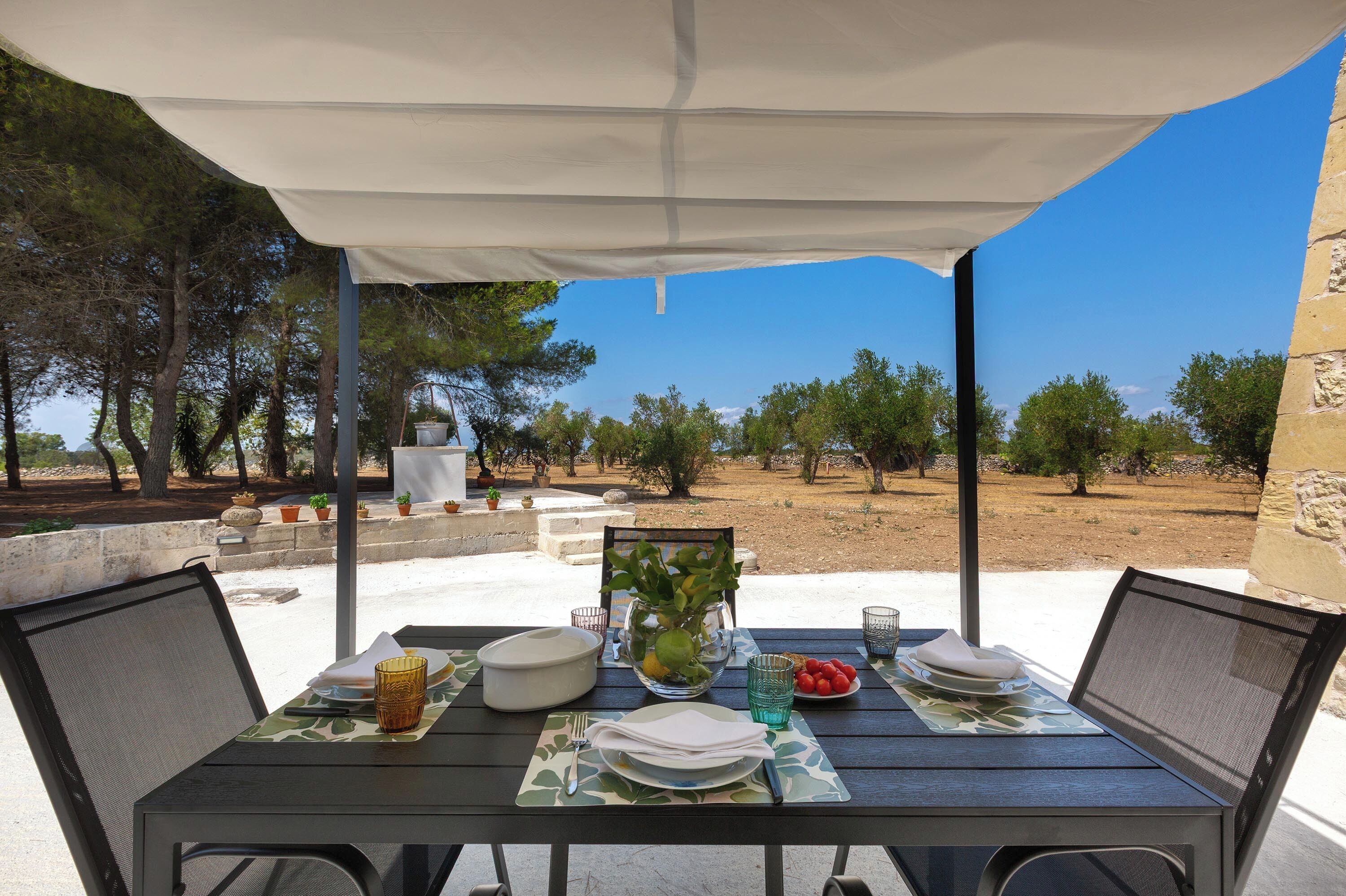Un área de comedor al aire libre con una mesa para dos, rodeada por un toldo blanco, con vista a un paisaje de árboles y cielo despejado.