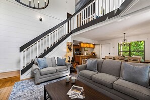 Un salon moderne avec un escalier, des canapés gris, une table basse avec des magazines et une cuisine en arrière-plan.
