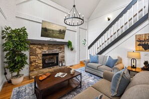 A spacious living room with a stone fireplace, a large plant, a grey sofa with blue cushions, and a staircase with a wooden railing. - New! Hot Tub/Game Room-Close to Dollywood (Pigeon Forge)