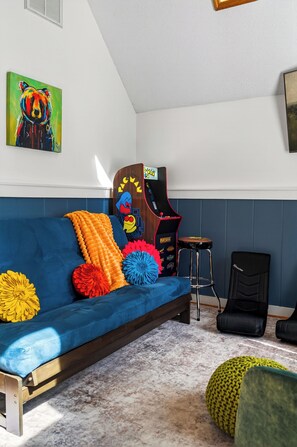 Ein Zimmer mit einem blauen Sofa, bunten Kissen, einer Pac-Man Spielmaschine und einem Gaming-Sessel.
