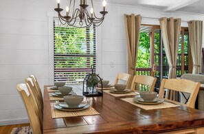 Une salle à manger avec une table en bois dressée pour quatre personnes, entourée de chaises, d’un lustre et d’une fenêtre avec des stores.