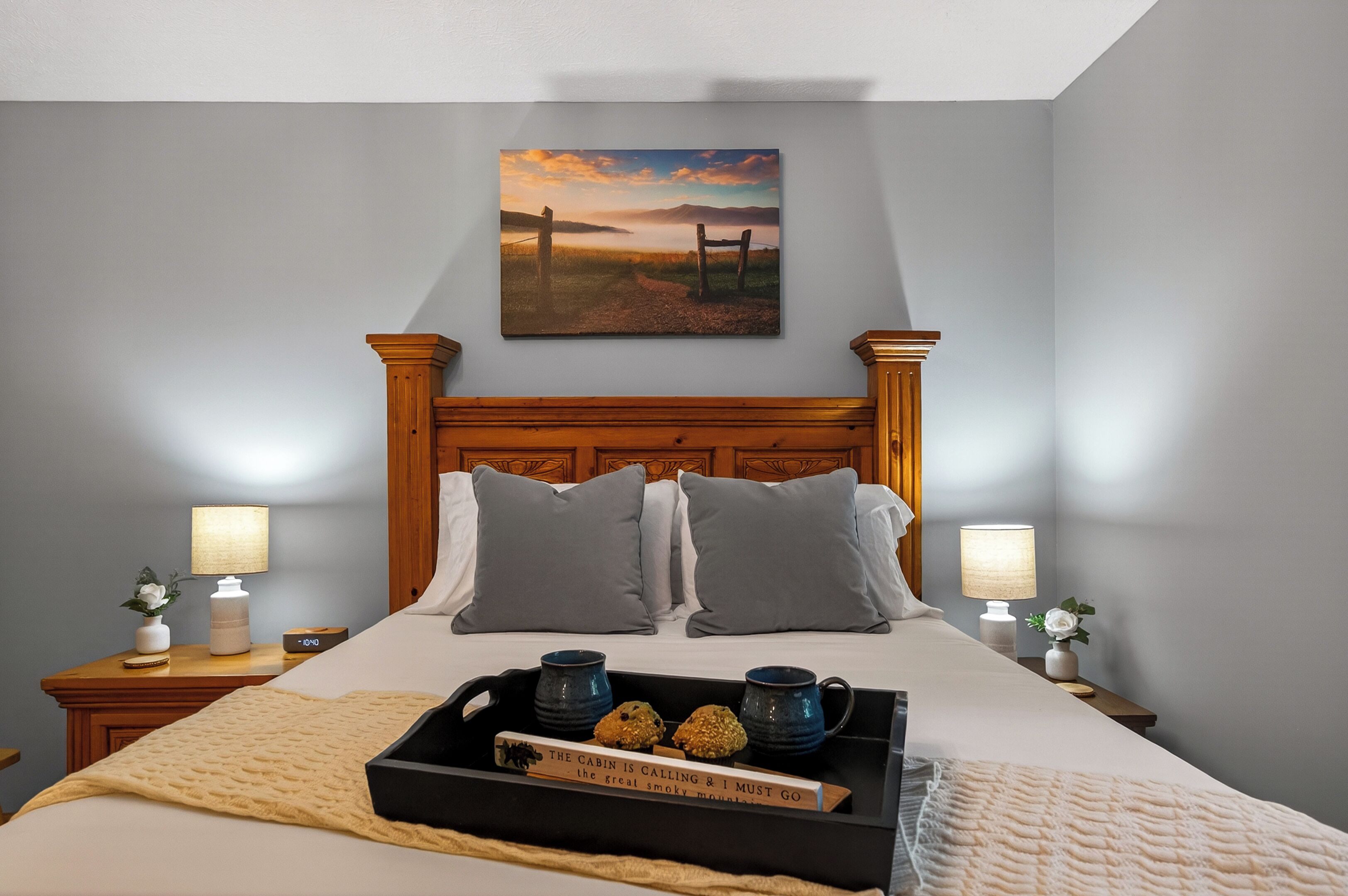 A bedroom with a wooden headboard, two bedside lamps, a painting of a landscape, and a tray with a book and a mug.