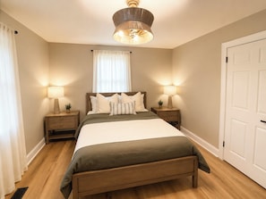 Un dormitorio con una cama, dos mesitas de noche, una lámpara de noche, una ventana con cortinas, una puerta y una lámpara en el techo.
