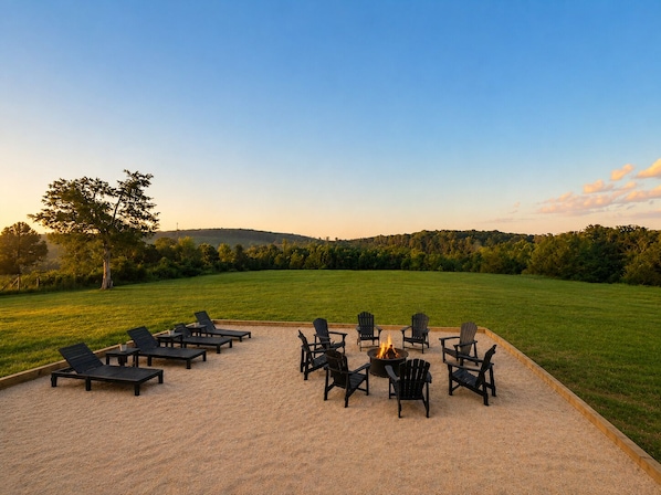 An outdoor seating area with lounge chairs and a fire pit, surrounded by a grassy field and trees.