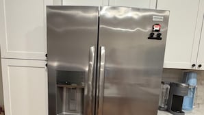 A stainless steel refrigerator with a water dispenser, located in a kitchen with white cabinets and a wooden floor.