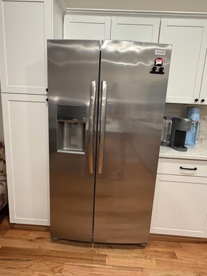 Un refrigerador de acero inoxidable con dispensador de agua, ubicado en una cocina con gabinetes blancos y piso de madera.
