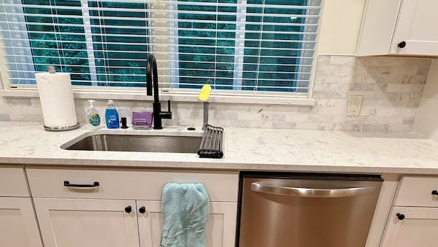 A kitchen with a stainless steel dishwasher, a sink with a faucet, and a window with blinds.