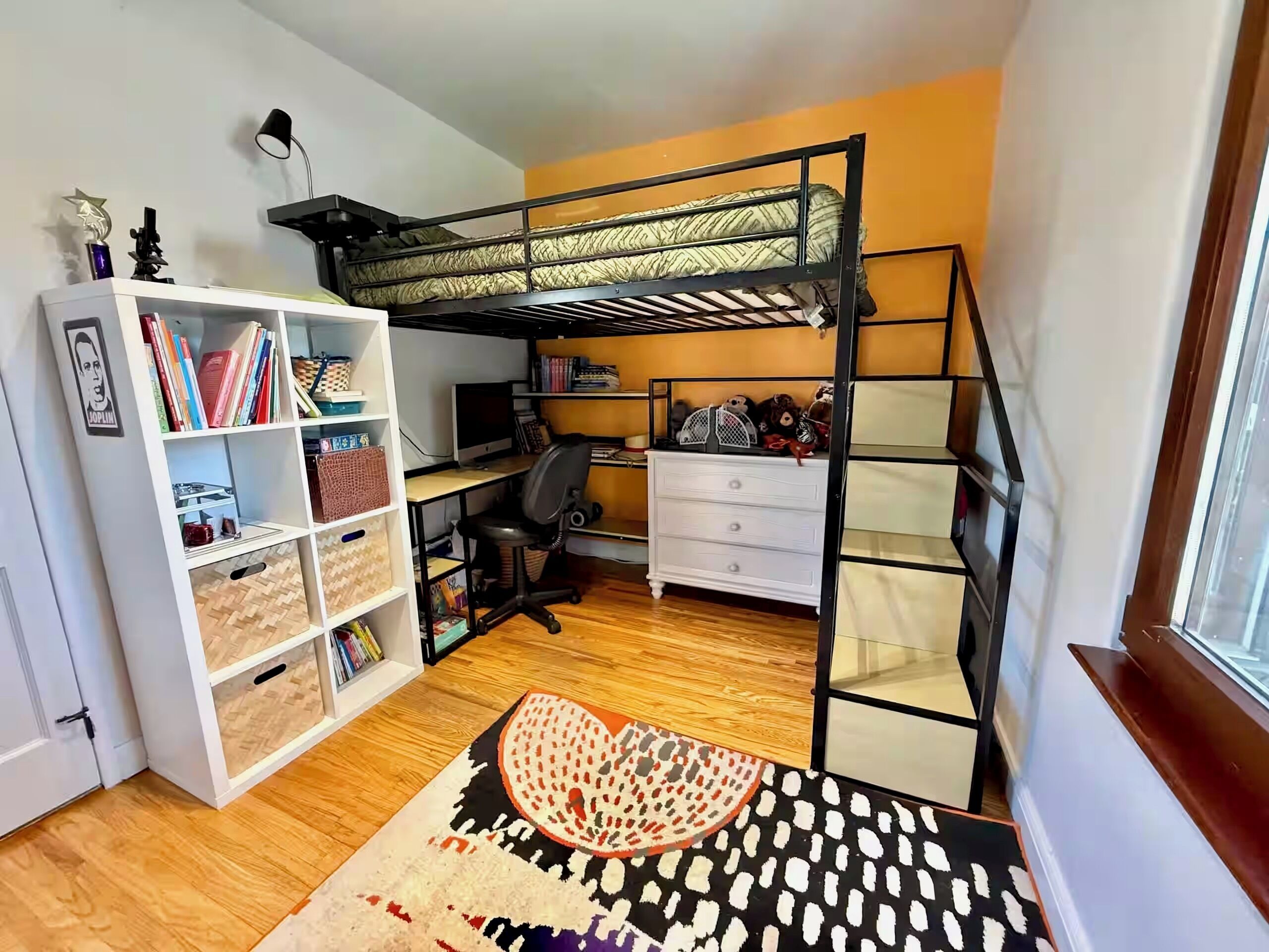 A compact room with a bunk bed, a desk with a computer, a bookshelf, and a patterned rug.