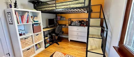 A compact room with a bunk bed, a desk with a computer, a bookshelf, and a patterned rug.