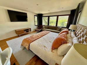 A modern bedroom with a large bed, a sofa, a TV mounted on the wall, and a window with a view of greenery. - Spanish Colonial Hilltop Retreat with 360° Views and Rooftop Deck (Los Angeles)