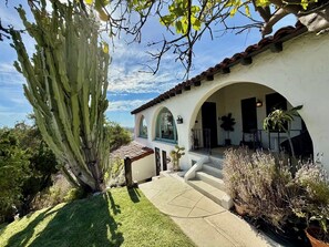 A white building with a tiled roof, an arched entrance, and a patio area with plants. - Spanish Colonial Hilltop Retreat with 360° Views and Rooftop Deck (Los Angeles)