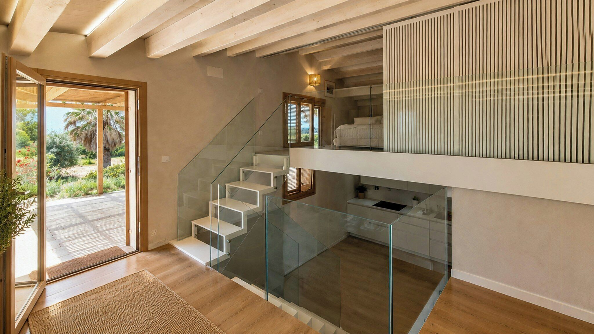 A modern interior with a glass staircase leading to a balcony, a kitchen area, and a view of a garden.