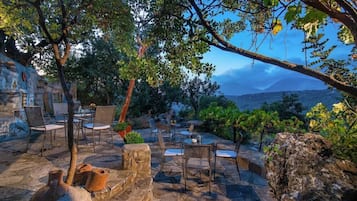 An outdoor seating area with stone tables and chairs, surrounded by trees and a view of distant mountains.