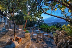 An outdoor seating area with stone tables and chairs, surrounded by trees and a view of distant mountains. - Enagron Ecotourism Village (Mylopotamos)