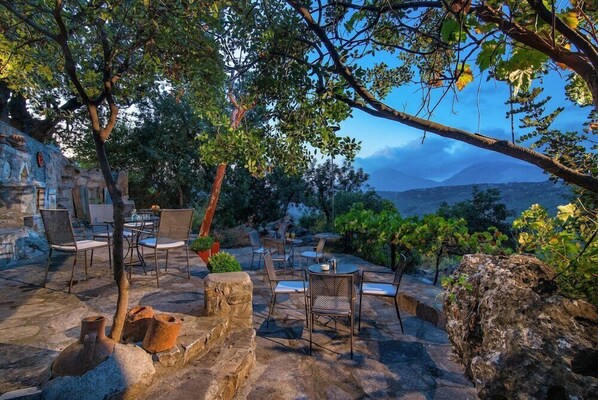 An outdoor seating area with stone tables and chairs, surrounded by trees and a view of distant mountains. - Enagron Ecotourism Village (Mylopotamos)