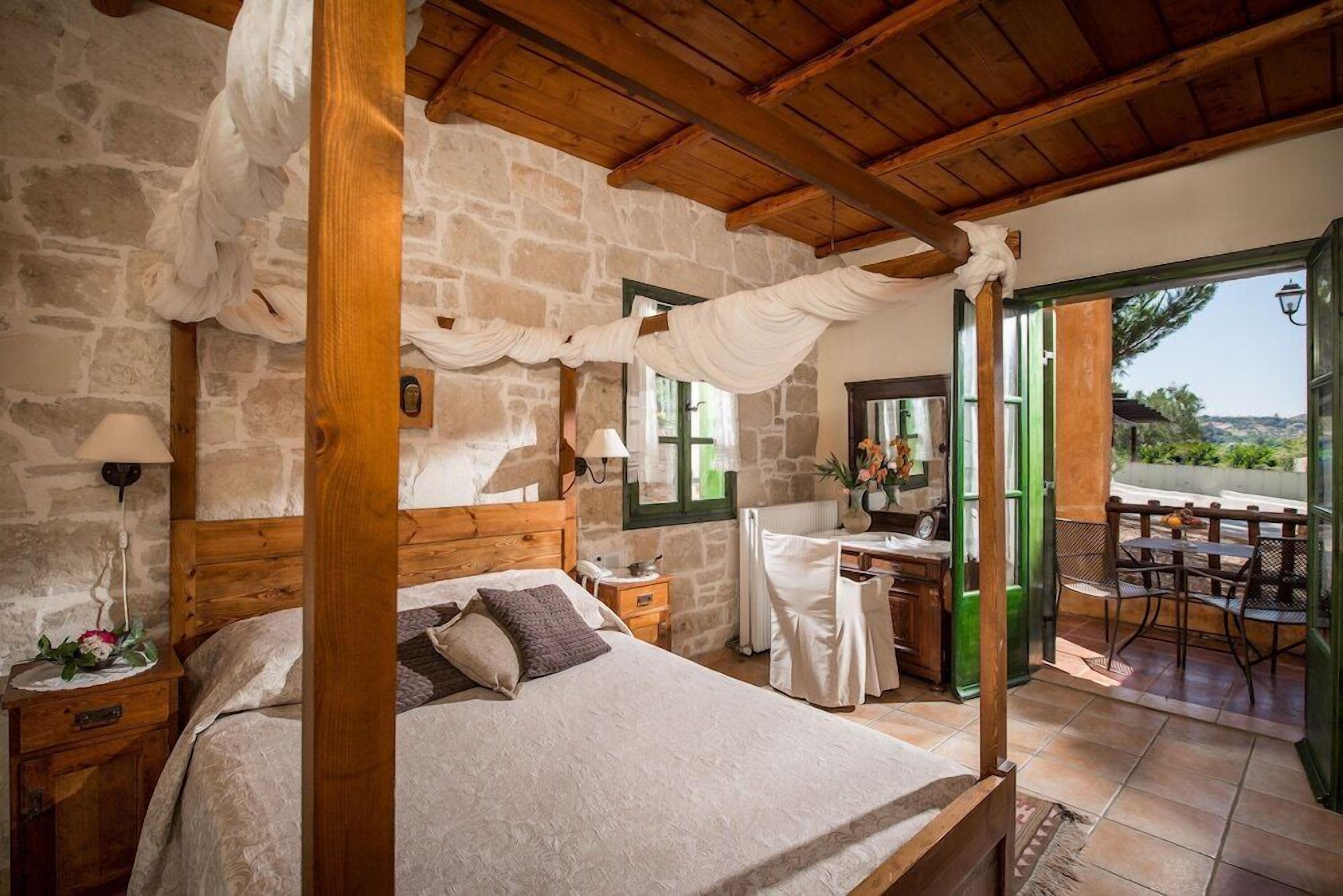 A bedroom with a stone wall, wooden ceiling beams, a bed with a canopy, a small table with a chair, and a view of the outdoors.