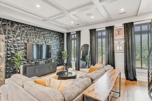 A modern living room with a sectional sofa, a flat-screen TV, a decorative wall, and large windows with curtains. - Pillar&Peak Modern NoMa 3BR| Rooftop Deck +Parking (Washington)