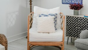 Ein Zimmer mit einem Korbstuhl, einer Lampe, einem an der Wand befestigten Schild mit der Aufschrift 'BEACH HOUSE' und einem gerahmten Bild.