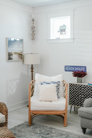 Ein Zimmer mit einem Korbstuhl, einer Lampe, einem an der Wand befestigten Schild mit der Aufschrift 'BEACH HOUSE' und einem gerahmten Bild.