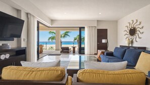 A modern living room with a flat-screen TV, a sofa, and a coffee table. There is a view of the beach and palm trees through an open door. - Beachfront 2BR Condo with Hot Tub at Diamante Dunes (Cabo San Lucas)