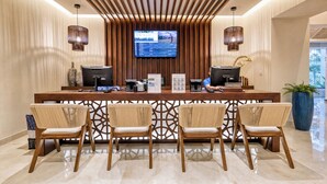 A reception desk with two monitors, a digital screen, and a concierge sign. There are four chairs with a woven backrest. - Beachfront 2BR Condo with Hot Tub at Diamante Dunes (Cabo San Lucas)