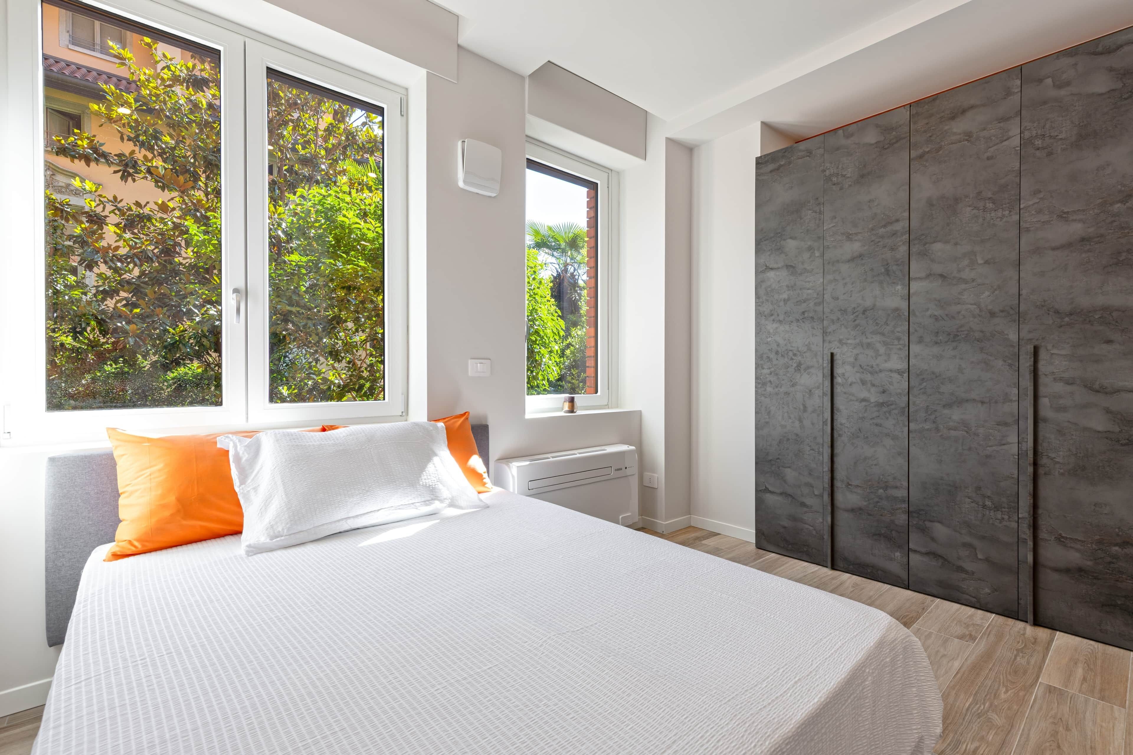 A modern bedroom with a large bed, two windows, a radiator, and a wardrobe.
