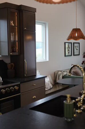 A modern kitchen with dark cabinetry, a stove, and a sink. There is a window providing natural light and a hanging pendant light. - Studio in Sugarcreek Steps away from Spa and Café (Sugarcreek)