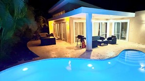 A swimming pool with a curved edge, a covered outdoor seating area with chairs and a table, and a building with large windows and a door. - Breezy, Beautiful 2 Bedroom Caribbean Villa! (Sosúa)