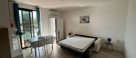 A bedroom with a bed, a desk, and a chair. There is a wardrobe and a small table with a lamp.