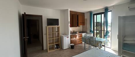 Une chambre avec un lit, un téléviseur posé sur un meuble en bois, un coin cuisine et une vue par la fenêtre.