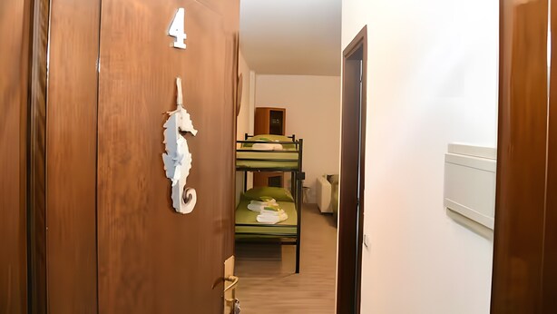 Un corridoio che porta a una stanza con letti a castello, una porta in legno con un numero e una parete bianca.