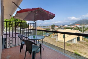A balcony with a table and chairs, a red umbrella, and a view of a town with buildings and a mountain in the distance.