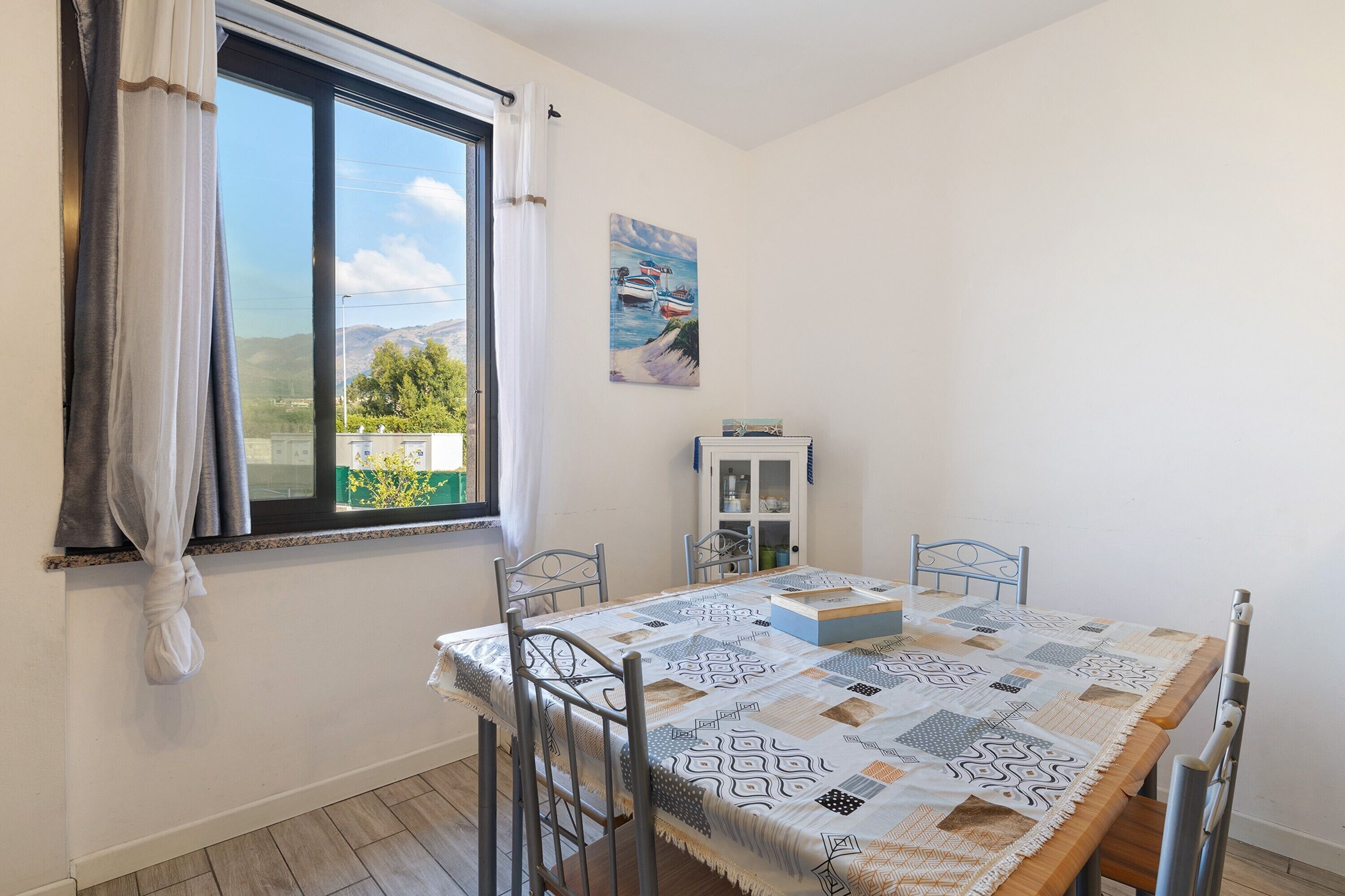 Un'area pranzo con tavolo apparecchiato per quattro persone, una finestra con tende e un quadro appeso al muro.