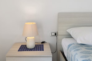 A bedside table with a lamp, a power outlet, and a bed with a patterned bedspread.
