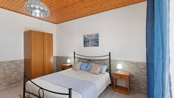 A bedroom with a bed, wooden wardrobe, nightstands, a wall-mounted lamp, and a painting.