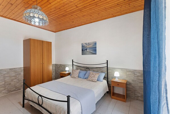 A bedroom with a bed, wooden wardrobe, nightstands, a wall-mounted lamp, and a painting.