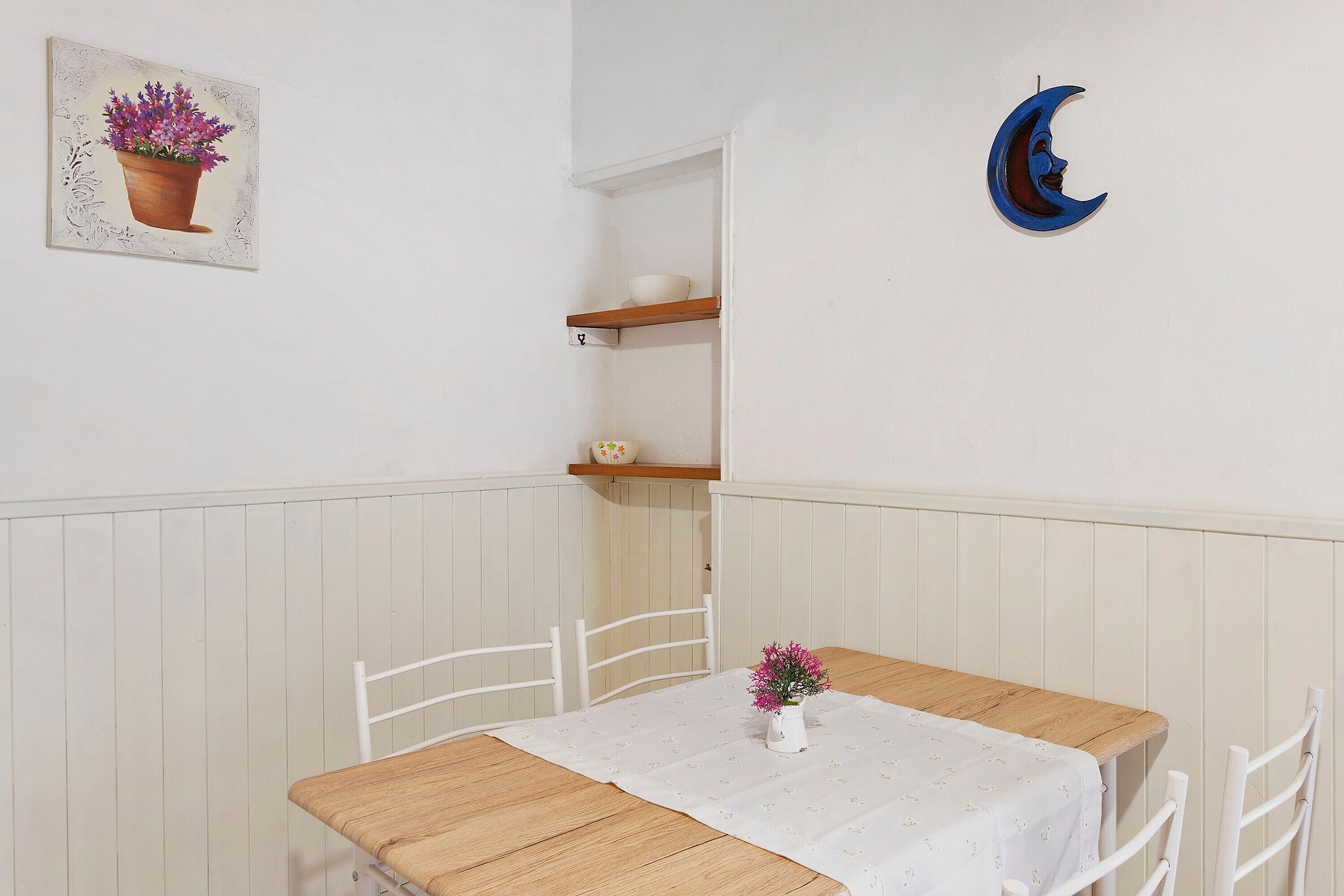En matplats med ett bord, stolar, en vit duk, en vas med blommor, en hylla med en skål och en halvmåneformad dekoration som hänger på väggen.
