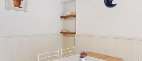 A dining area with a table, chairs, a white tablecloth, a vase with flowers, a shelf with a bowl, and a wall-mounted crescent moon decoration.