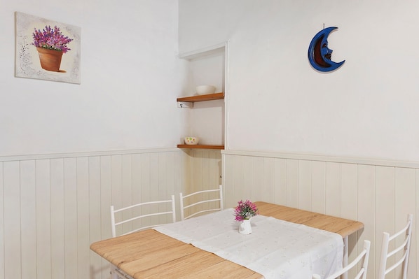 A dining area with a table, chairs, a white tablecloth, a vase with flowers, a shelf with a bowl, and a wall-mounted crescent moon decoration.