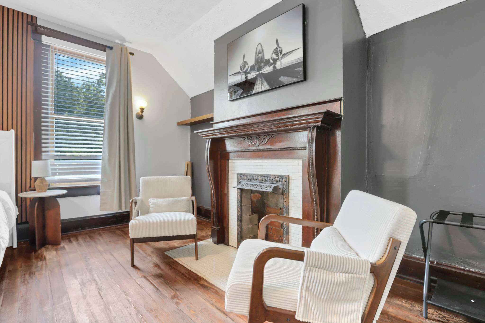 A room with a fireplace, two upholstered chairs, a lamp, and a painting on the wall.