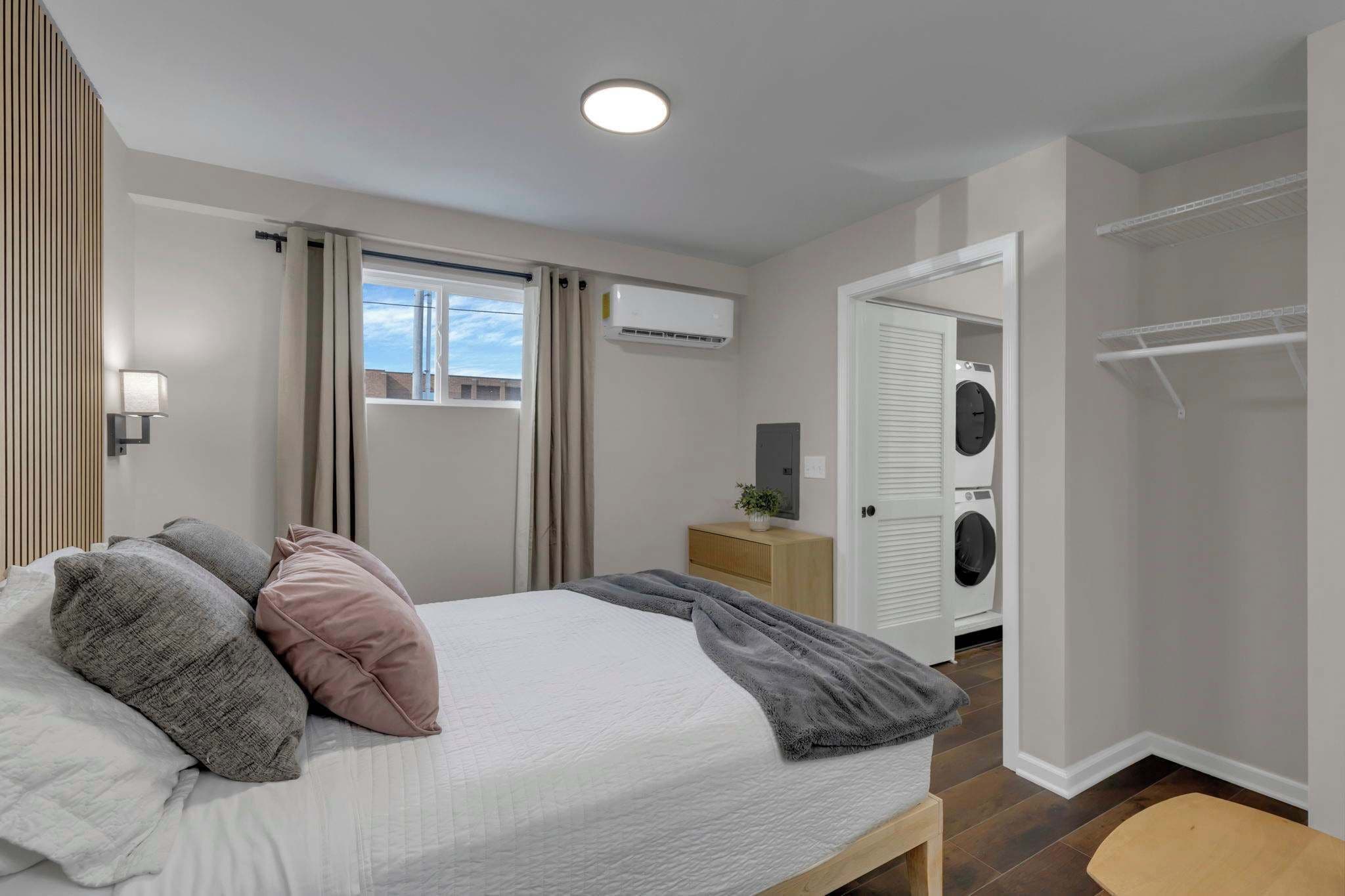 A bedroom with a bed, a bedside table, an air conditioning unit, a window with curtains, a washing machine, and a closet.