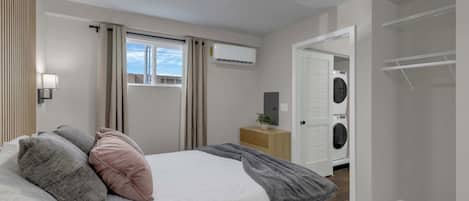Un dormitorio con cama, mesita de noche, aire acondicionado, ventana con cortinas, lavadora y armario.