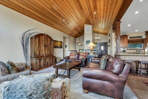 A spacious living room with a high wooden ceiling, a fireplace, leather sofas, and a dining area with a kitchen in the background. - Tahoe City Retreat | Lake View & Hot Tub (Tahoe City)