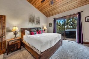 A bedroom with a large bed, wooden headboard, two bedside tables with lamps, a wall-mounted artwork, and a large window with curtains. - Tahoe City Retreat | Lake View & Hot Tub (Tahoe City)