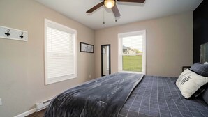 Un dormitorio con cama, ventilador de techo, dos ventanas con persianas y vista a un jardín verde.