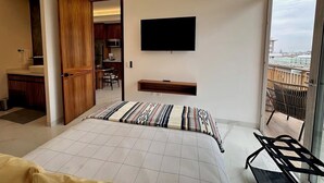 Ein modernes Schlafzimmer mit Bett, Wand-Fernseher, Balkon mit Aussicht und einer durchgehenden Tür erkennbaren Küche.