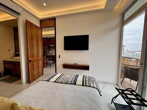 A modern bedroom with a bed, a TV mounted on the wall, a balcony with a view, and a kitchen visible through an open door. - Luxury & Modern Penthouse in the Heart of Zona Romántica - Walk Everywhere! (Puerto Vallarta)