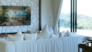 Ein Zimmer mit Steinwand, einem großen Gemälde einer Strandansicht, einem weißen Sofa mit vielen Kissen und Blick auf eine bewaldete Landschaft durch ein Fenster.
