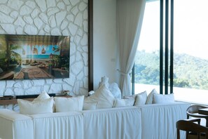 A room with a stone wall, a large painting of a beach scene, a white sofa with multiple cushions, and a view of a forested area through a window. - Luxury 6-Bedroom Villa with Sea View and Gym (Chaweng)